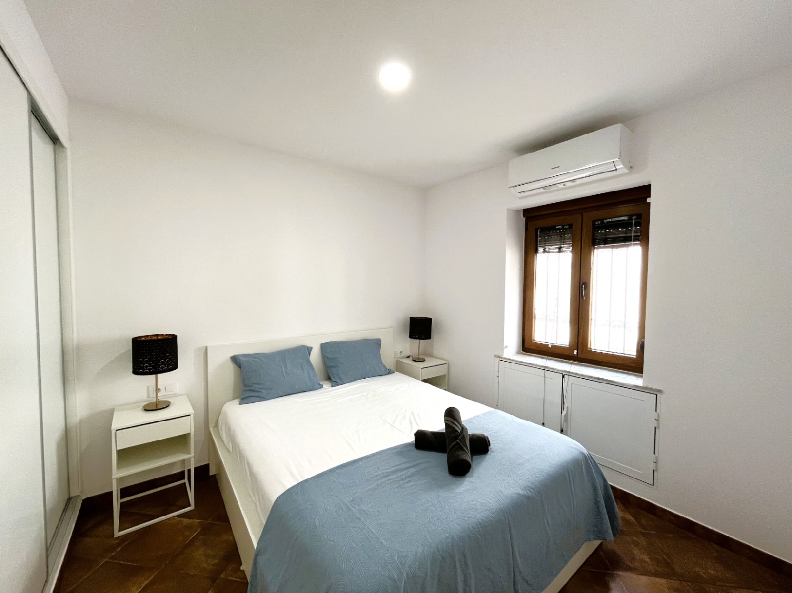 Slaapkamer met tweepersoonsbed, witte nachtkastjes en airconditioning. Voorzien van een rustieke tegelvloer, inbouwkast en houten raam met rolluiken.