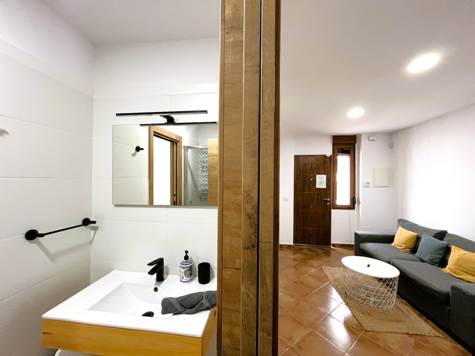 Woonkamer met terracotta vloer en grijze bank, grenzend aan een moderne badkamer met wit wasmeubel, zwarte kranen en strakke witte betegeling.