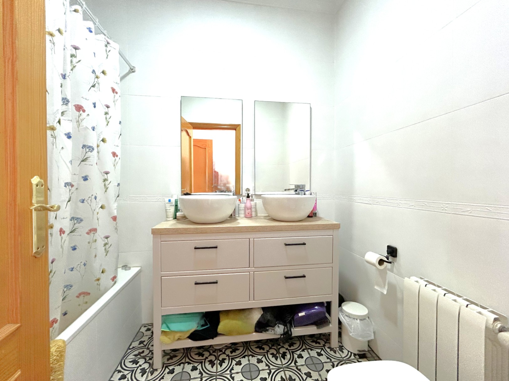Badkamer met dubbele wastafel op houten blad, decoratieve hydraulische vloertegels, ligbad en radiator voor centrale verwarming.