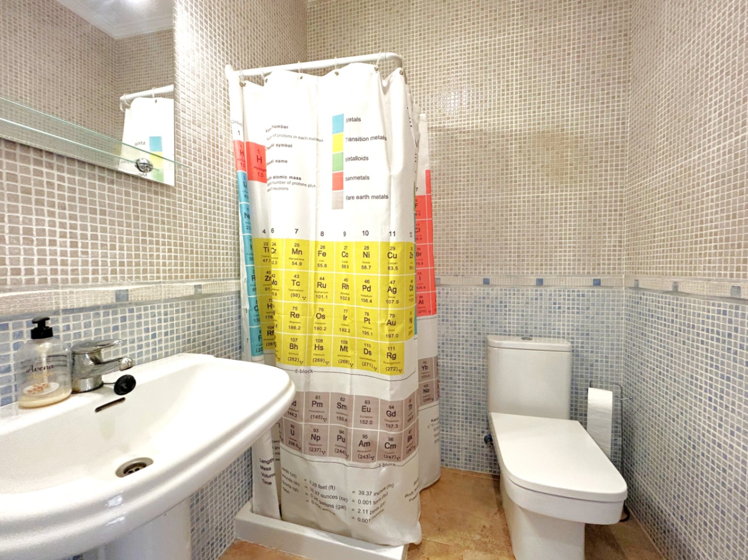 Badkamer met tweekleurige mozaïekwandtegels, een witte wastafel op zuil en een douchegordijn met het periodiek systeem. Voorzien van een modern toilet en spiegel met glazen planchet.