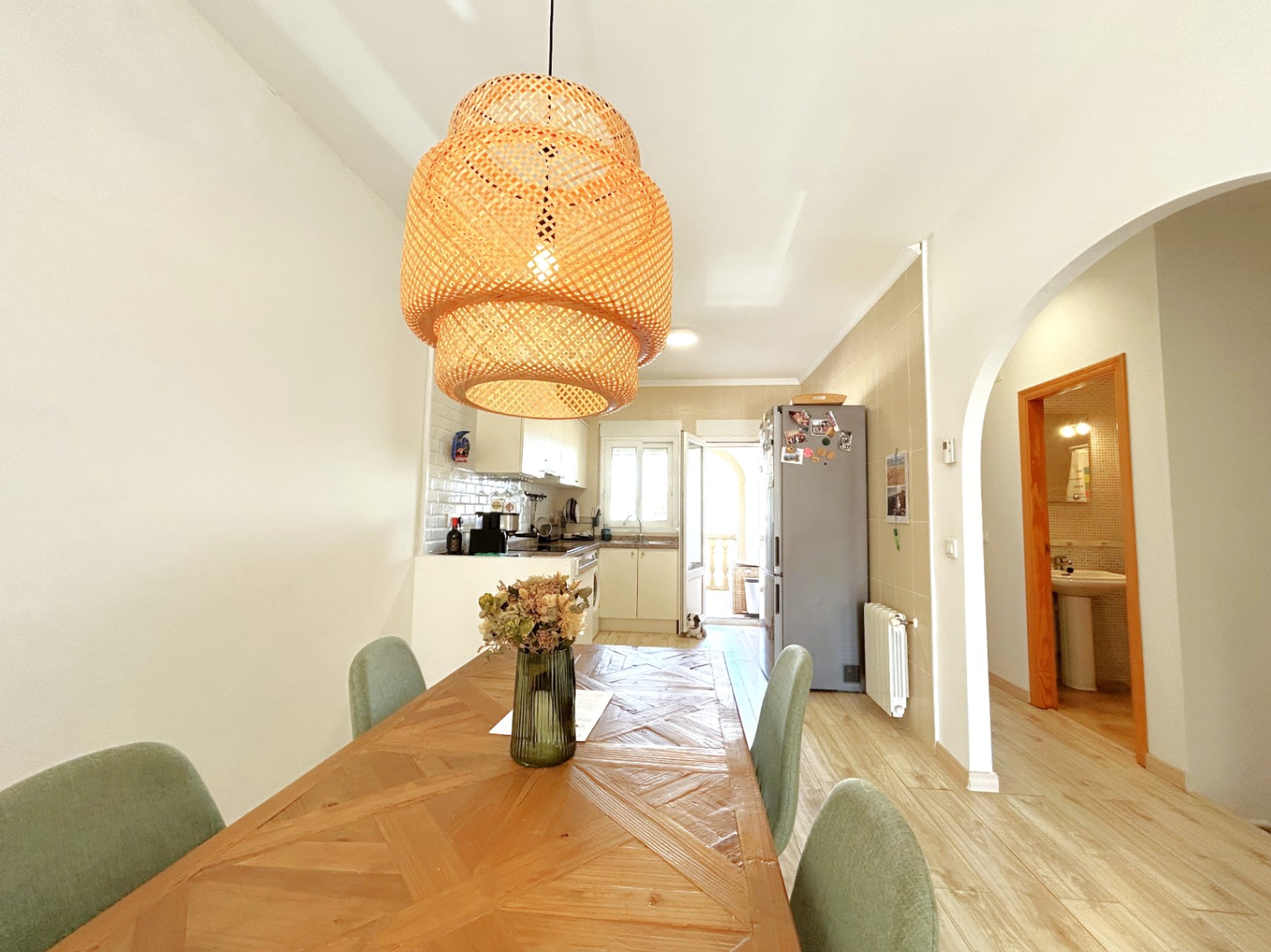 Eethoek met geometrische houten tafel en rotan hanglamp, grenzend aan een open witte keuken met doorgang naar de praktische bijkeuken.