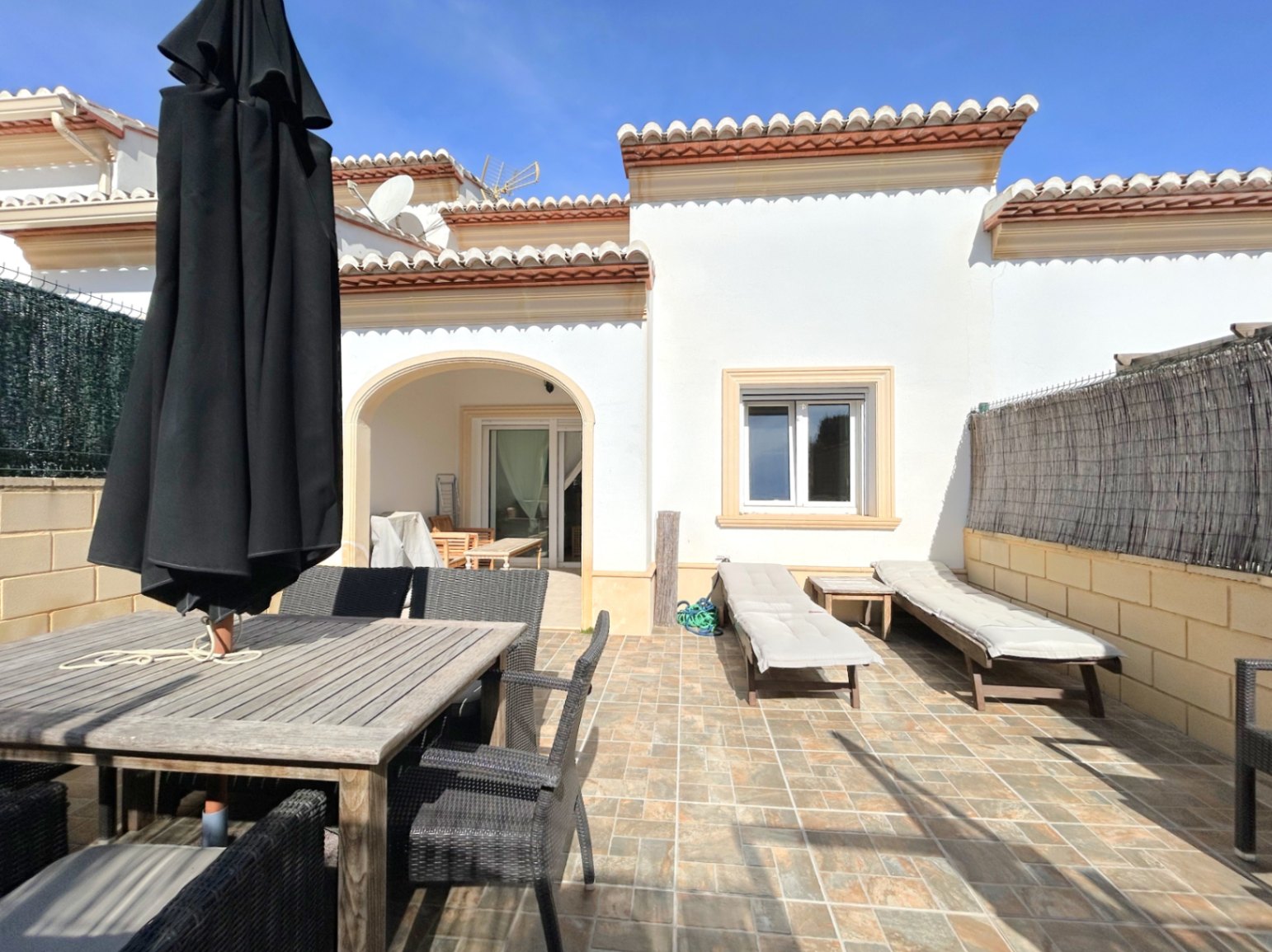 Privéterras met tegelvloer, houten eethoek en ligbedden. De mediterrane gevel is voorzien van een overdekt terras met boog en traditionele dakpannen.