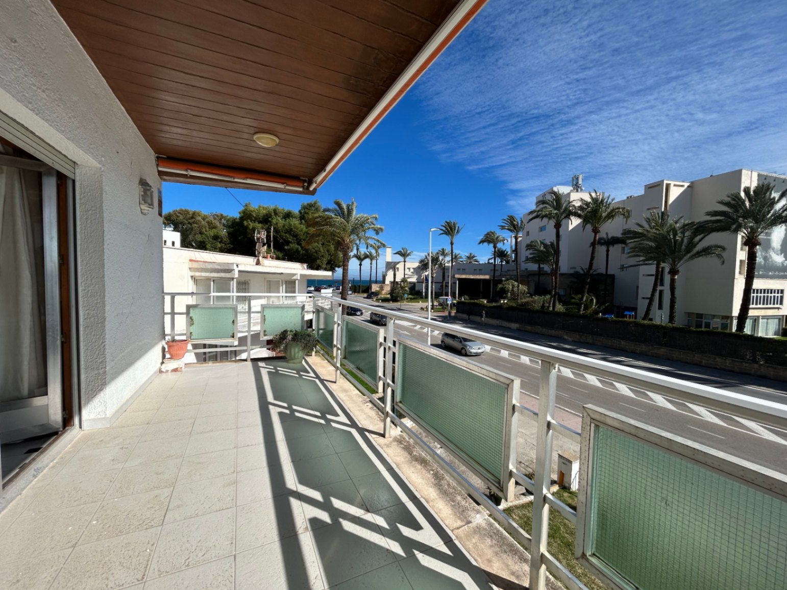 Buitenterras met keramische tegels, een houten plafondafwerking en metalen balustrades met glas. Het biedt uitzicht op een laan met palmbomen.