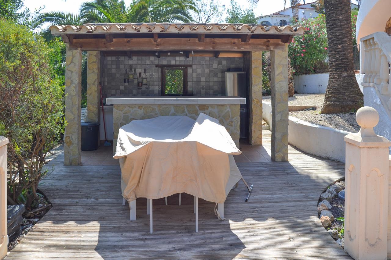 Buitenkeuken met een met steen beklede bar, gemetselde pilaren en een pannendak op een houten terras, omgeven door mediterrane beplanting.