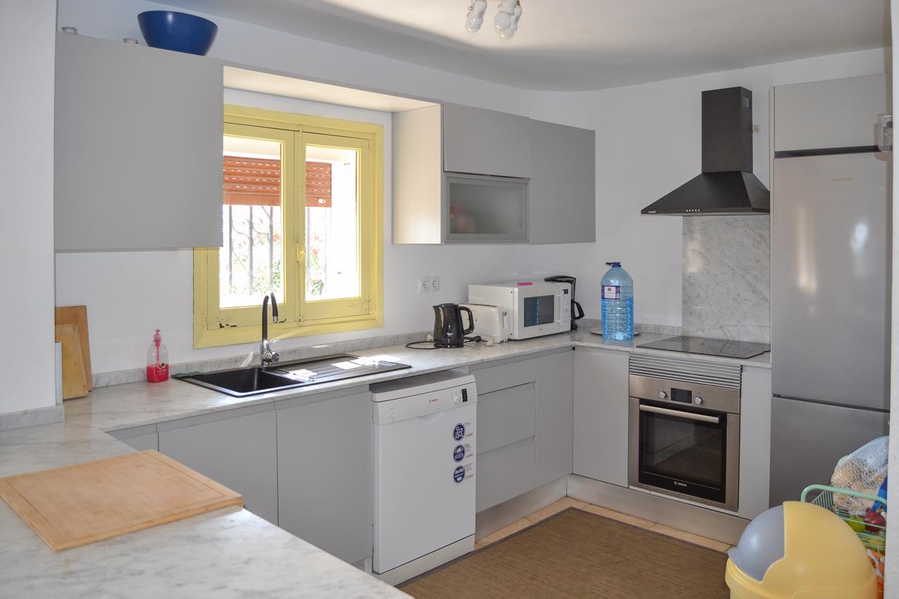 Functionele keuken met grijze kasten, marmerlook werkblad en inbouwapparatuur. Voorzien van een vaatwasser, oven en een raam met geel kozijn voor natuurlijk licht.