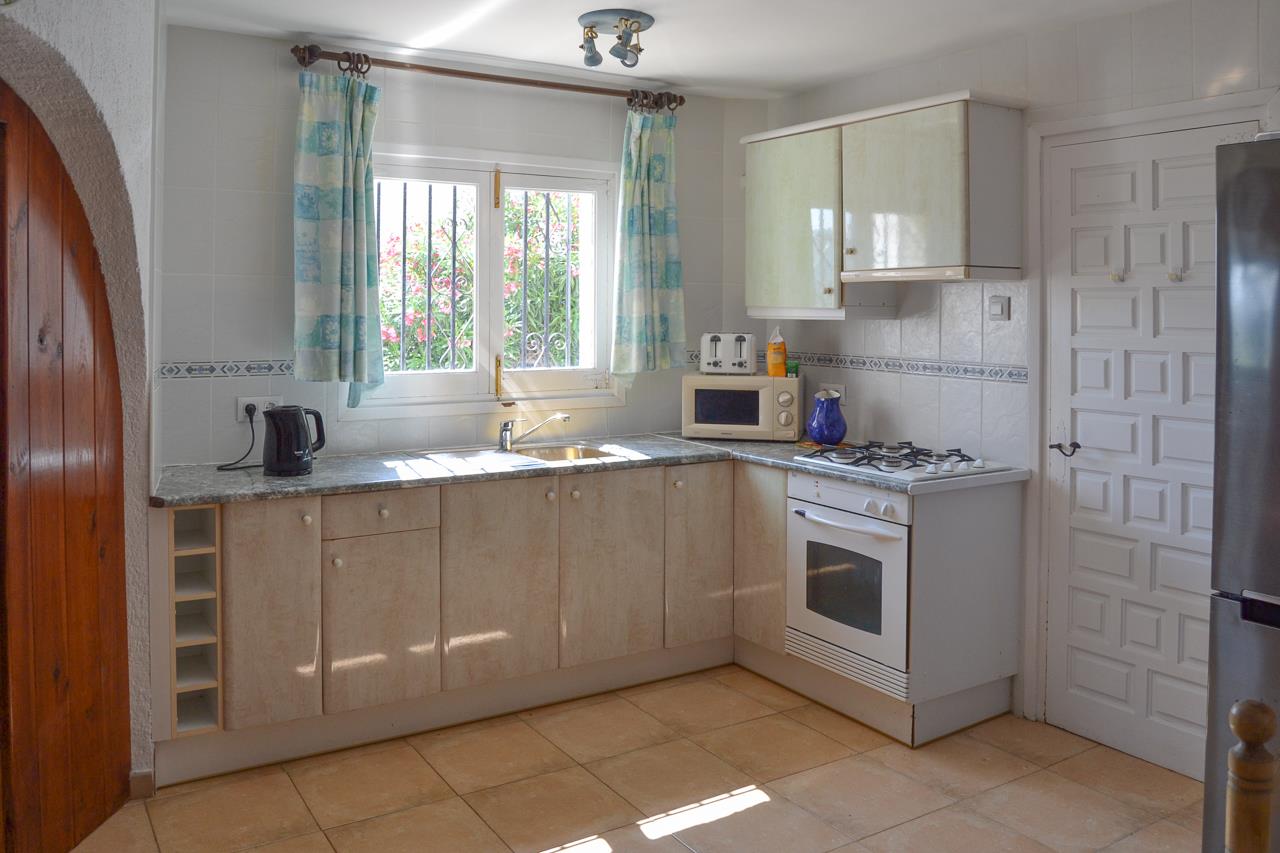 Functionele keuken met werkblad in granietlook, witte oven en gaskookplaat. Een raam met veiligheidstralies biedt natuurlijk licht en uitzicht op de tuin.