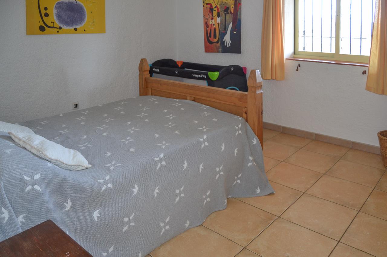 Lichte slaapkamer met houten tweepersoonsbed, reiswieg en tegelvloer. De kamer heeft een raam met tralies, gele gordijnen en kleurrijke schilderijen aan de wand.