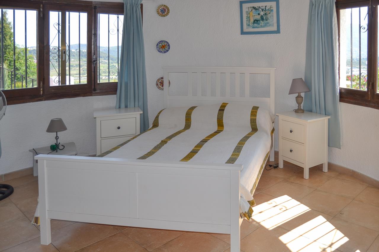 Lichte slaapkamer met wit houten bed, terracotta tegelvloer en ramen met donkerhouten kozijnen die uitzicht bieden op de vallei van Monte Pedreguer.