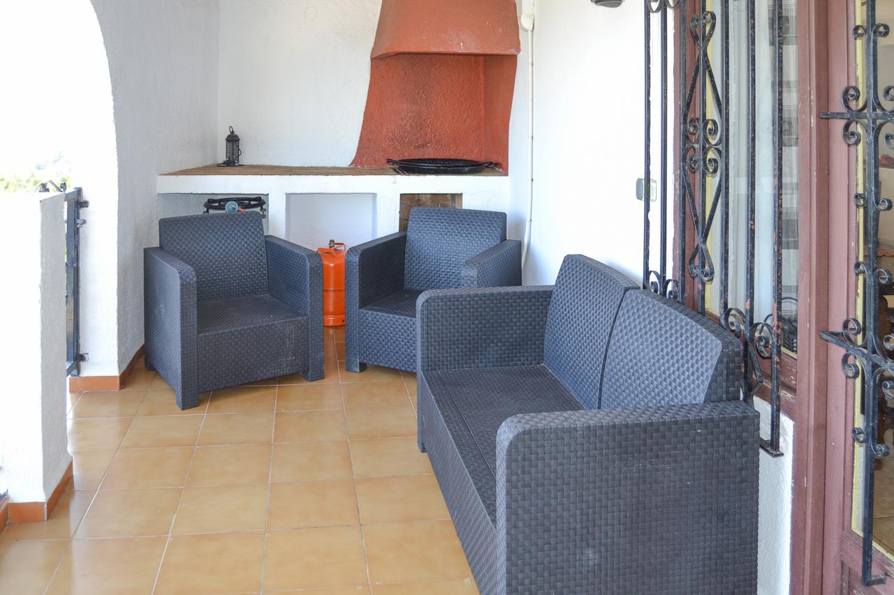 Overdekt terras met donker synthetisch rotan meubilair, terracotta vloertegels en een ingebouwde barbecue met een karakteristieke schoorsteenkap.