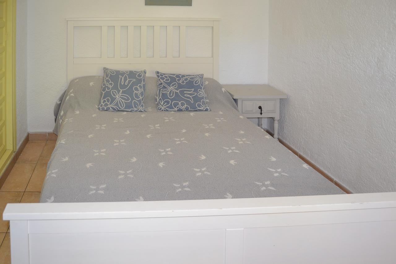 Slaapkamer met een wit houten bedframe, lattenbodem hoofdbord en bijpassend nachtkastje. Voorzien van een keramische tegelvloer en witte structuurwanden.