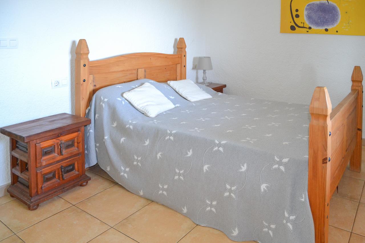 Slaapkamer met massief houten bedframe, rustiek nachtkastje en een tegelvloer. De witte muren met textuur vormen een contrast met het traditionele houten meubilair.