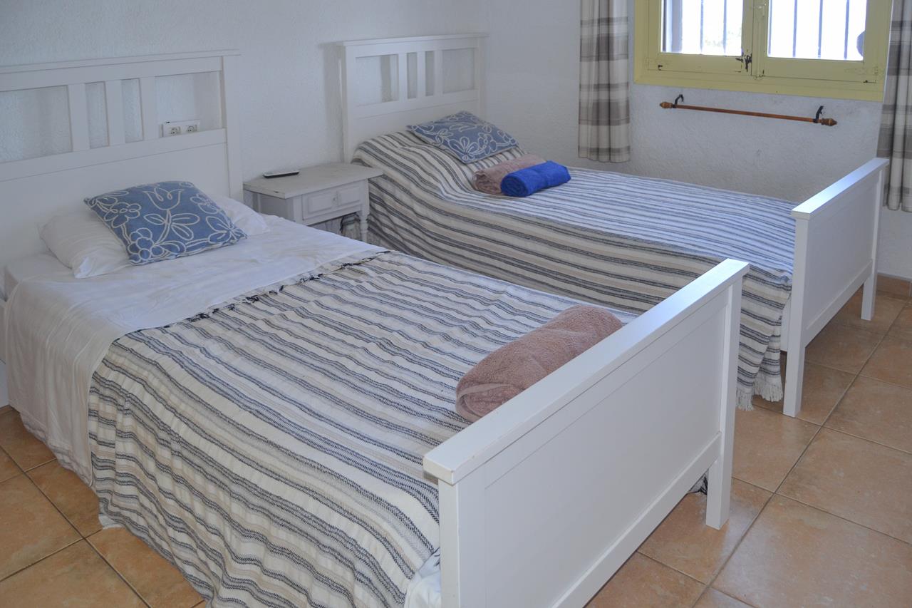 Slaapkamer met twee wit houten eenpersoonsbedden, rustieke tegelvloer en raam met geel kozijn en geruite gordijnen.
