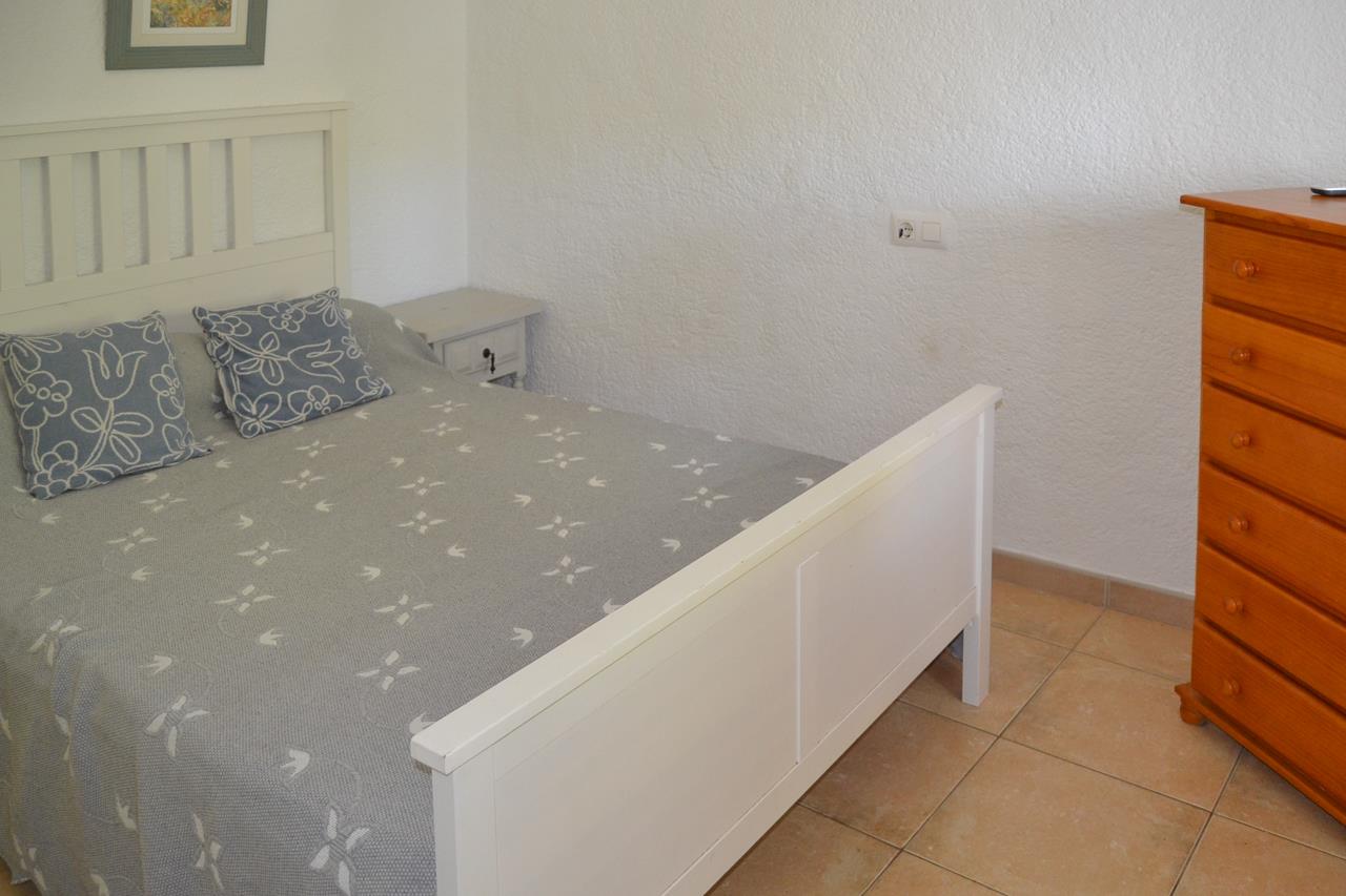 Slaapkamer met wit houten bedframe, keramische tegelvloer en een houten ladekast. De witte muren met textuur versterken de natuurlijke lichtinval.