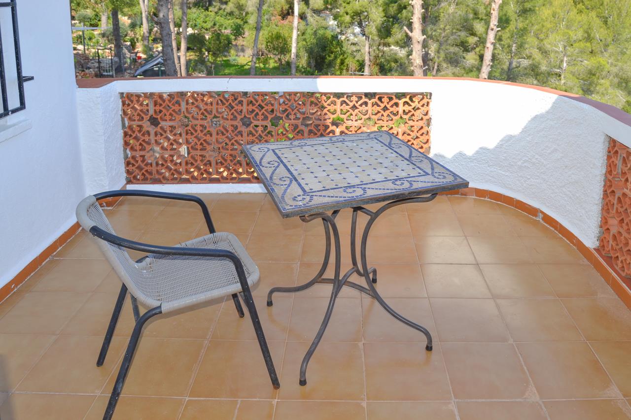 Terras met terracotta tegels, een tafel met blauw mozaïekblad en een decoratieve keramische balustrade met uitzicht op het omliggende dennenbos.