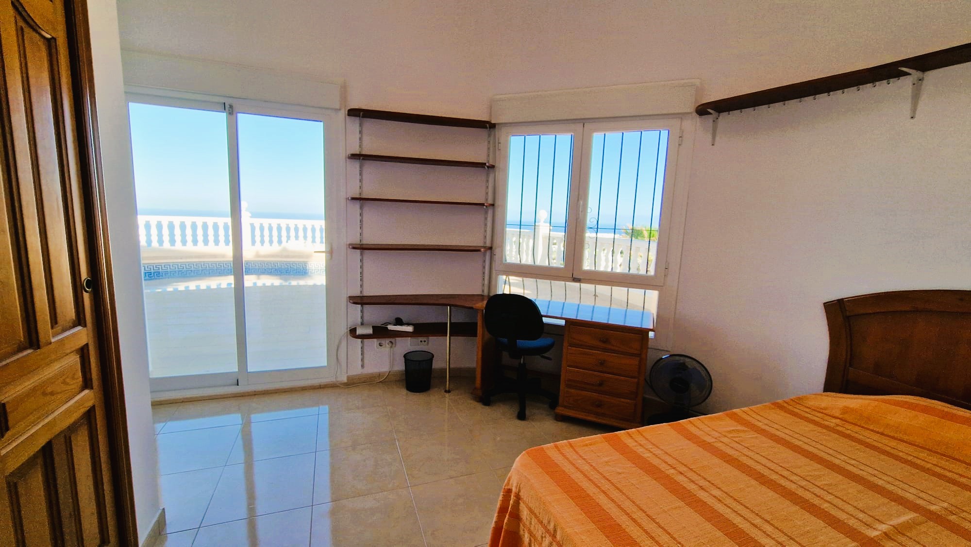 Lichte slaapkamer met houten bureau, planken en uitzicht op het balkon met witte balustrade.