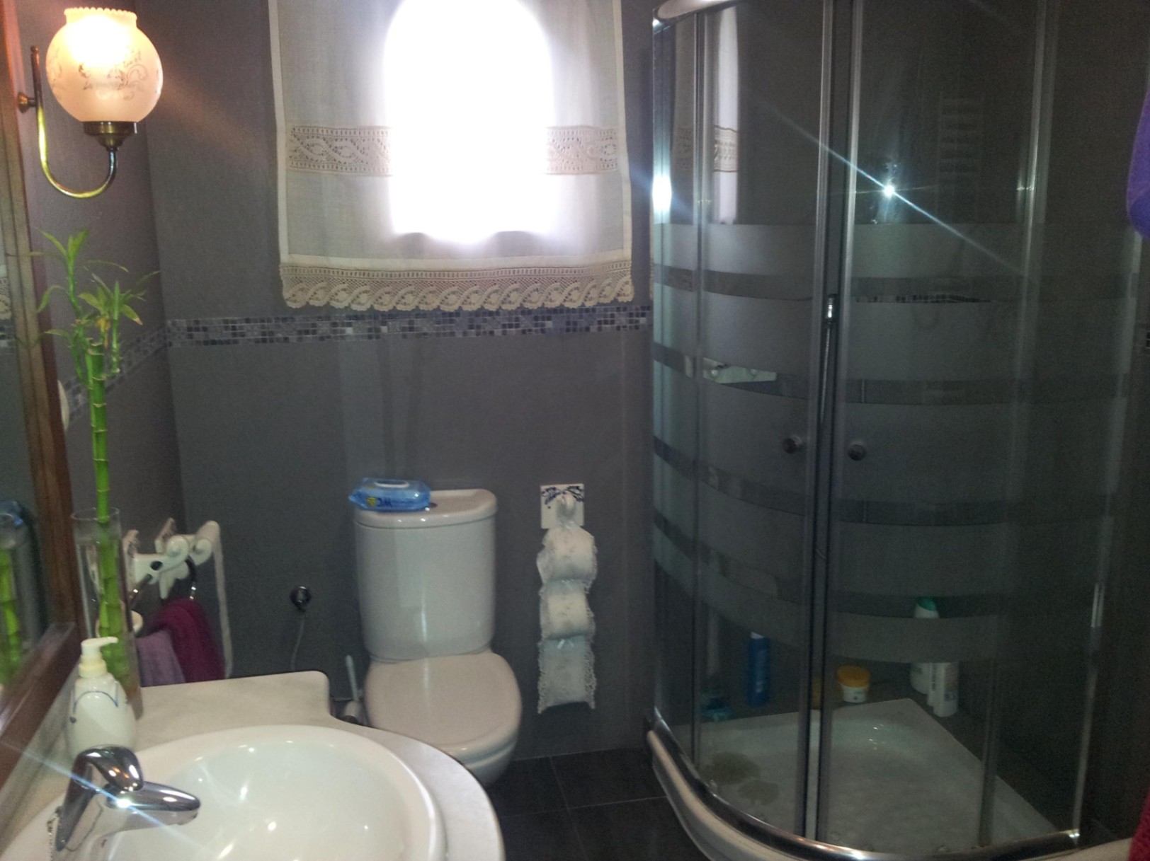 Badkamer met een gebogen glazen hoekdouche, grijze tegelwanden met mozaïekrand en een witte wastafel.