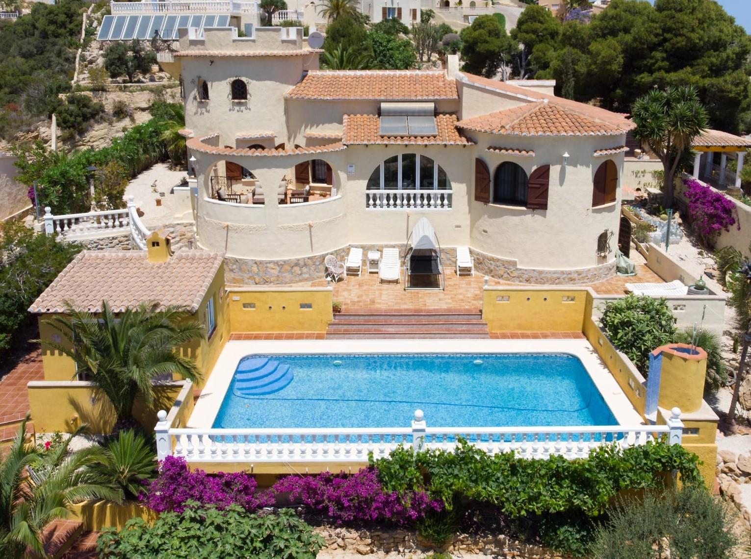 Buitenaanzicht van de villa met rechthoekig zwembad, terracotta terras en een kenmerkende toren, omgeven door mediterrane beplanting en gele muren.