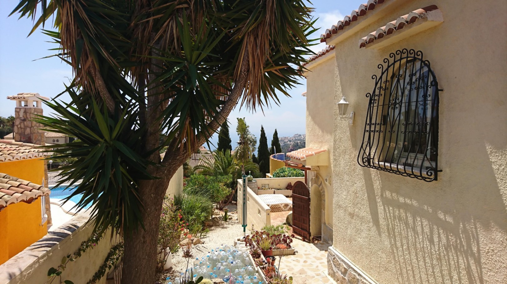 Exterieur van de villa met gestucte muren, smeedijzeren raamrooster en mediterrane tuin met yucca's en uitzicht op zee in de verte.