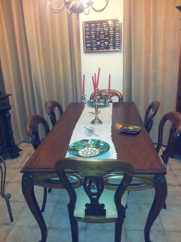 Formele eetkamer met een donkerhouten tafel, bewerkte stoelen en kamerhoge beige gordijnen. Gedecoreerd met zilveren kandelaars en mozaïekborden.