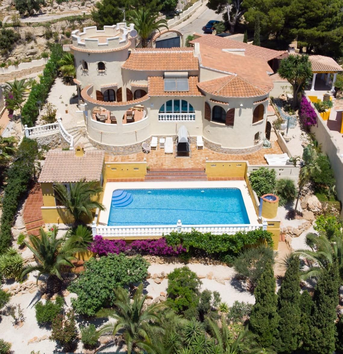 Luchtfoto van de mediterrane villa met ronde toren, een zwembad van 10x4m en een volgroeide tuin met palmbomen en kleurrijke bougainville op een ruim perceel.