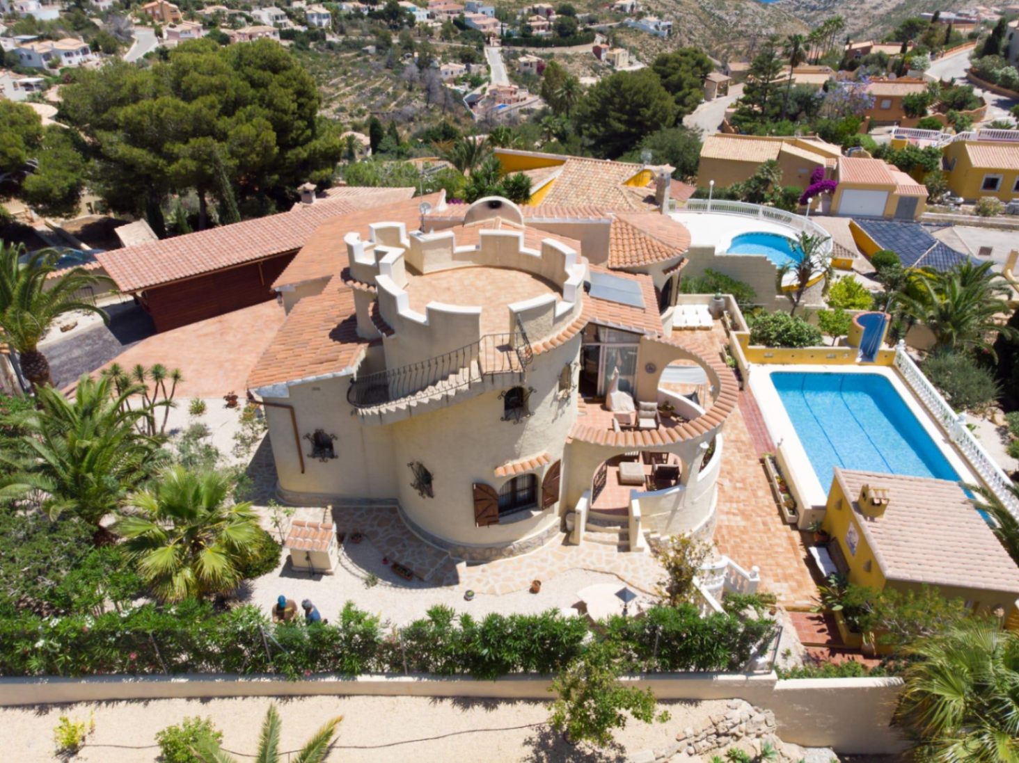 Luchtfoto van de villa met een karakteristieke ronde toren, dakterras, rechthoekig zwembad en buitenkeuken te midden van een mediterrane tuin.