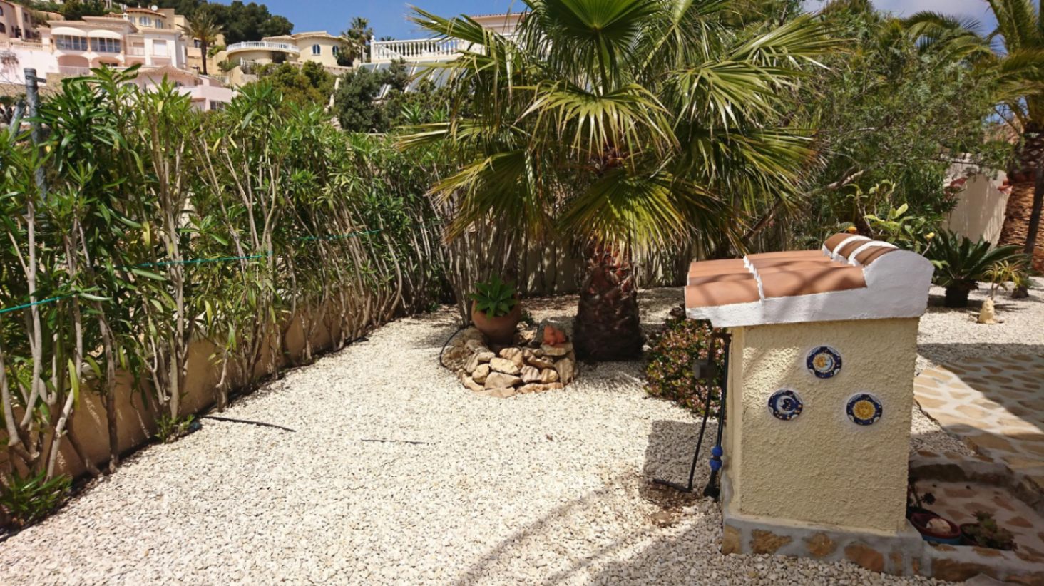 Mediterrane tuin met wit grind, een palmboom en een decoratief element met traditionele keramische tegels en een pannendak.