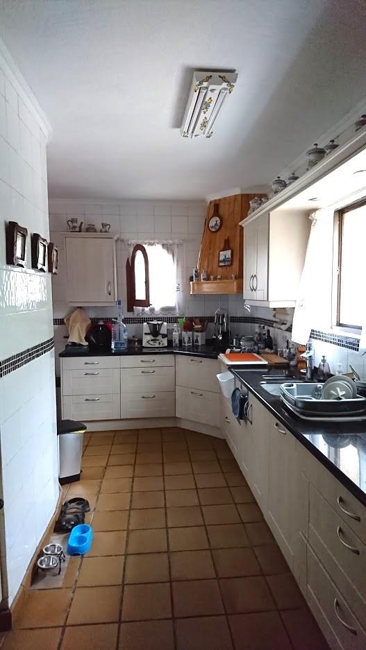 Onafhankelijke keuken met witte kasten, donkere werkbladen en een terracotta tegelvloer. Voorzien van een houten afzuigkap en natuurlijk licht via de ramen.
