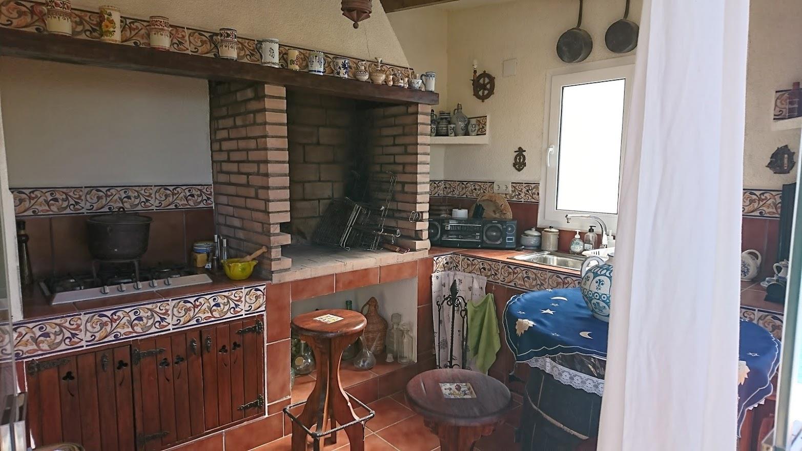 Rustieke buitenkeuken met een ingebouwde bakstenen barbecue, traditionele tegels, houten kasten en een spoelbak bij het raam.