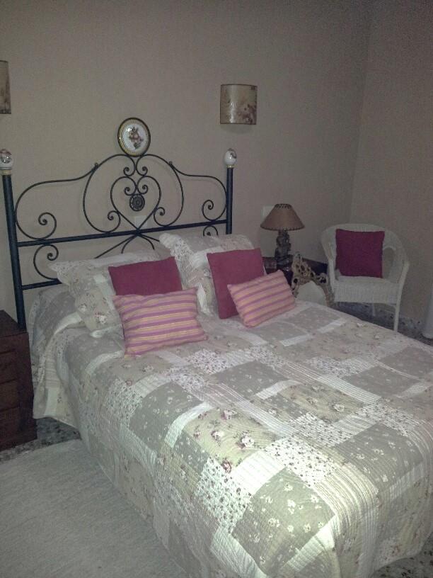 Slaapkamer met een smeedijzeren hoofdbord, patchwork sprei en een witte rieten stoel. Voorzien van wandarmaturen en een klassieke schemerlamp naast het bed.