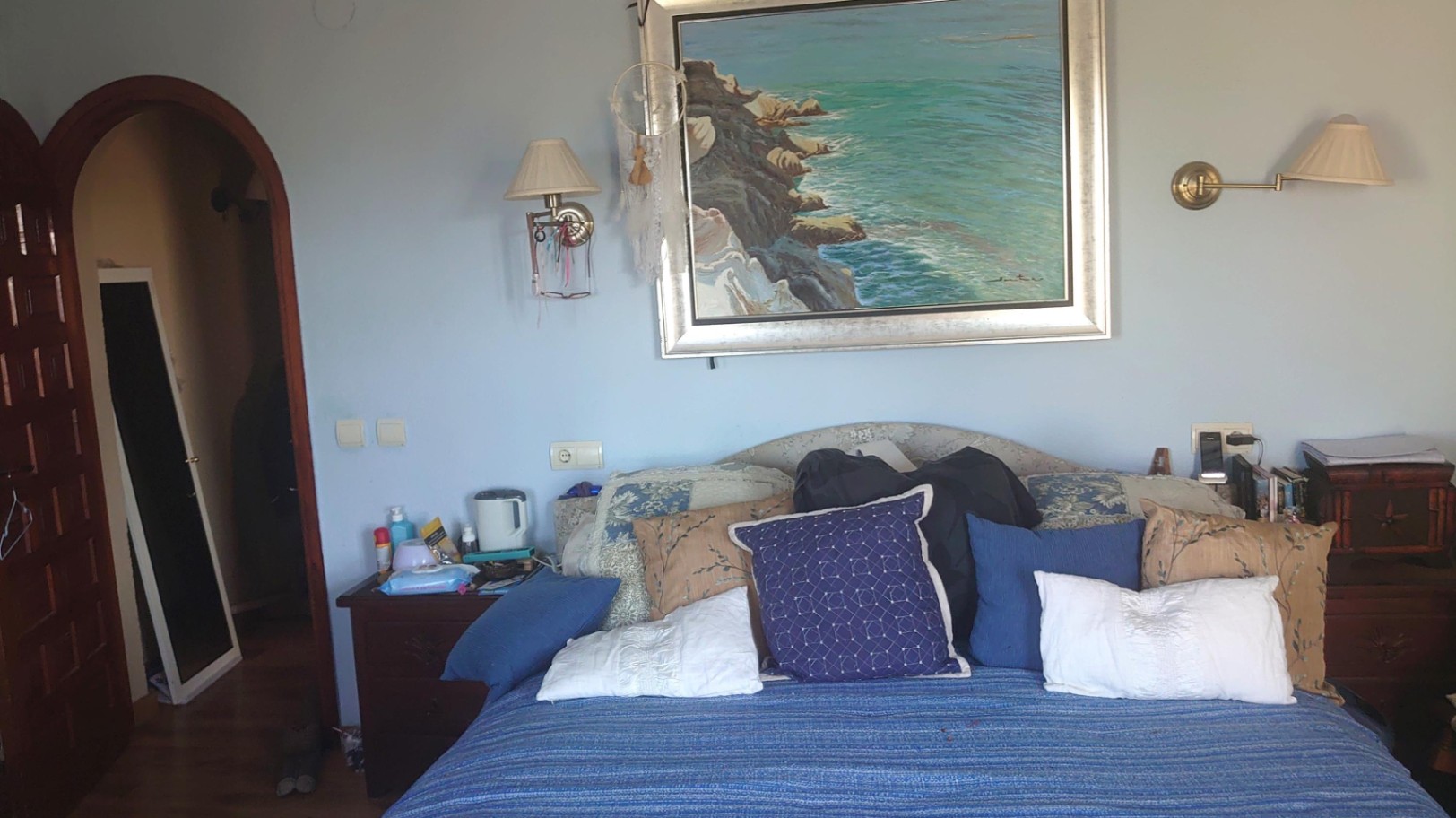 Slaapkamer met lichtblauwe muren, een groot schilderij van de kust boven het bed, wandlampen en een karakteristieke houten toogdeur.