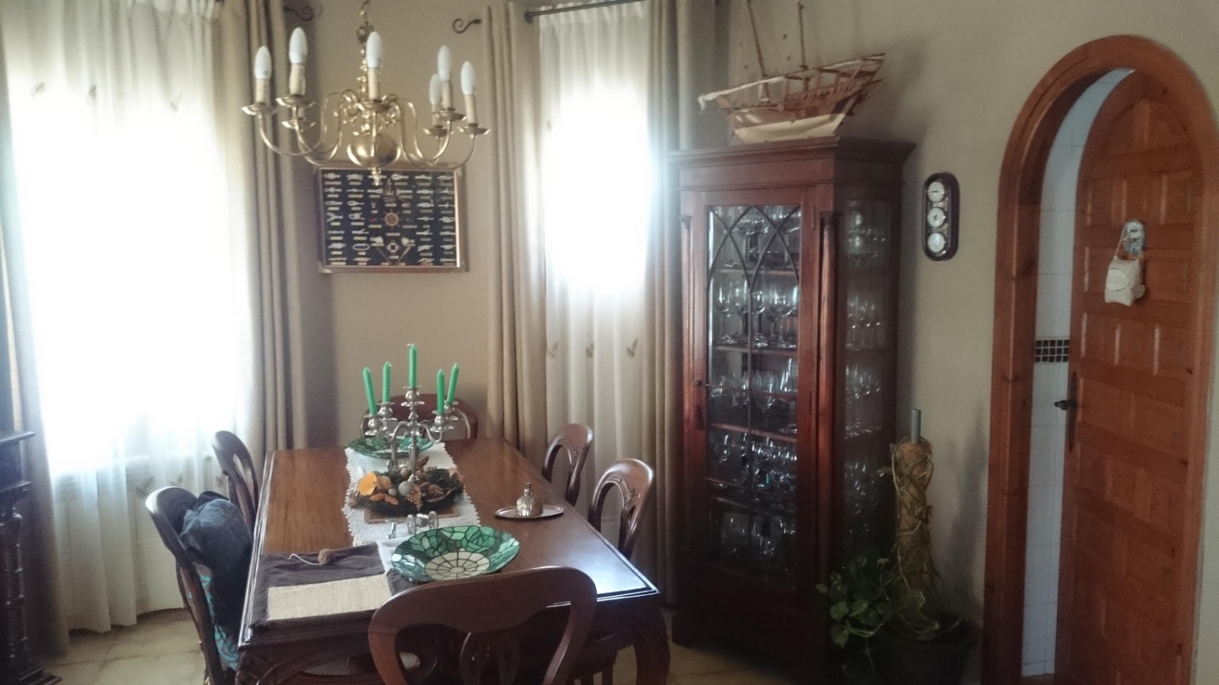Traditionele eetkamer met houten meubilair, een vitrinekast en koperen kroonluchter. De ruimte is voorzien van een houten boogdeur en natuurlijk licht.