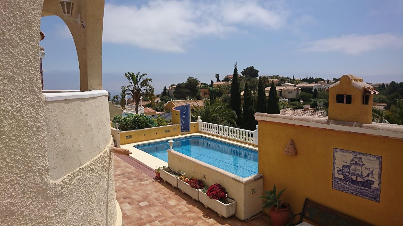 Verhoogd zicht op het rechthoekige zwembad met terracotta terras, okerkleurige muren met decoratieve tegels en mediterrane tuin met uitzicht op zee.