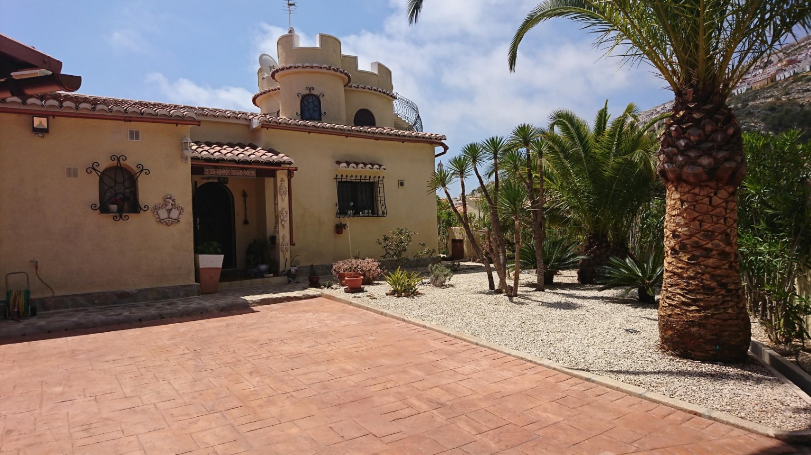 Vooraanzicht van de villa met een karakteristieke ronde toren, terracotta dakpannen en een mediterrane grindtuin met volwassen palmbomen.