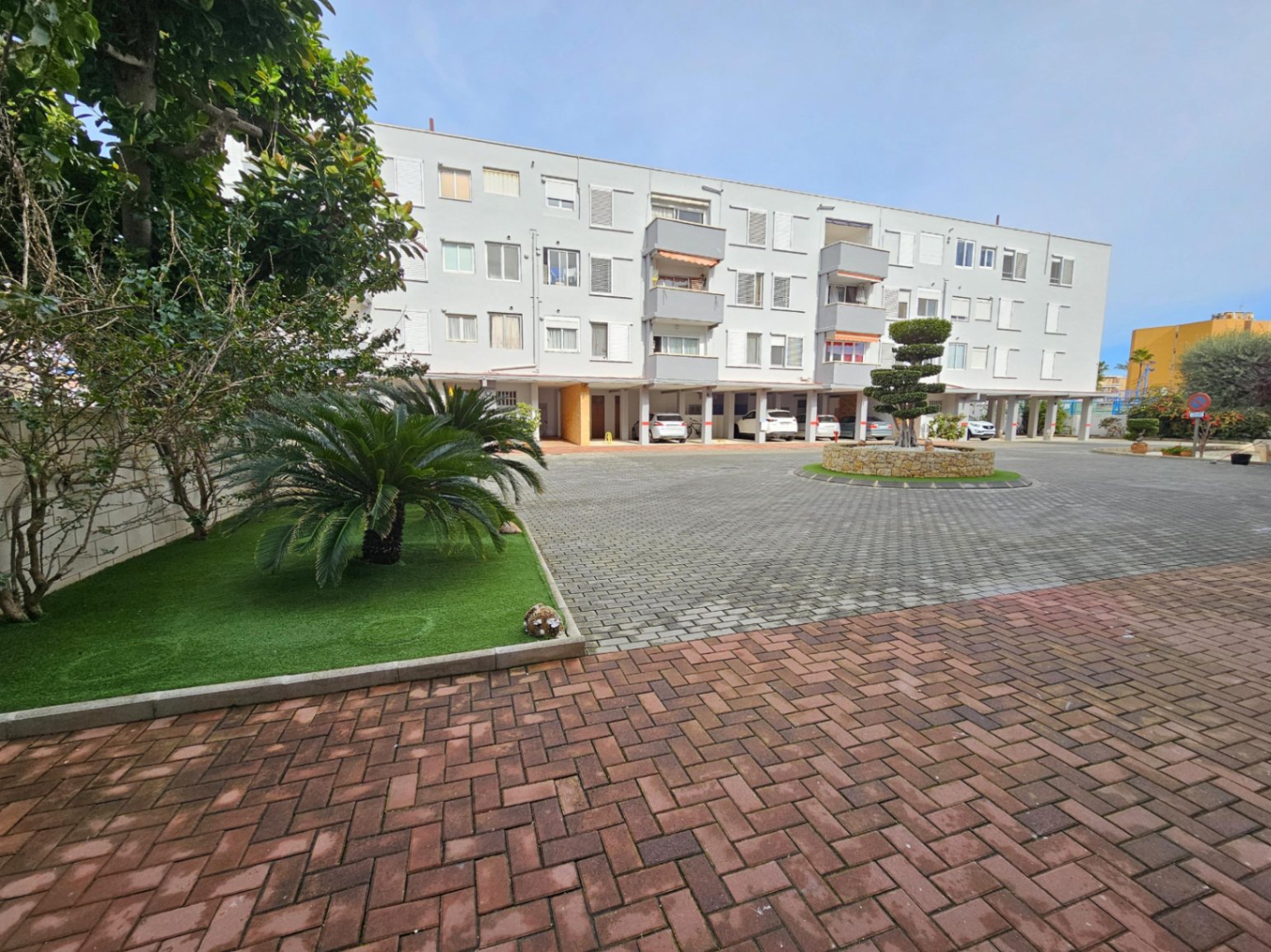 Buitenaanzicht van het wooncomplex met een geplaveide binnenplaats, aangelegde tuinzones en overdekte parkeerplaatsen op de begane grond.