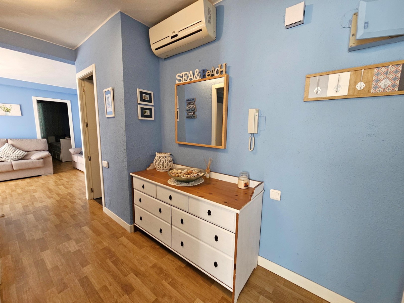 Entreehal met blauwe muren, wit houten dressoir, parketvloer en een split-airco-unit, met doorkijk naar de lichte woonkamer.