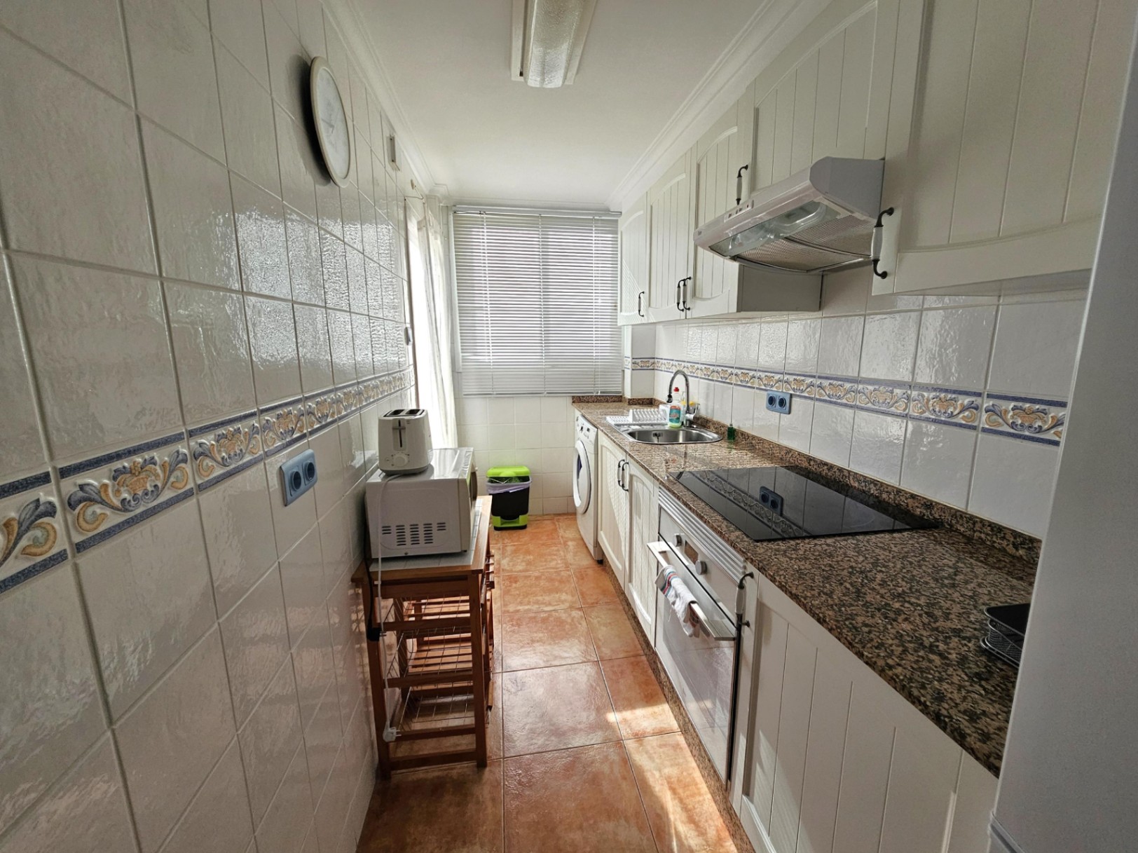 Gerenoveerde langwerpige keuken met granieten werkblad, witte kasten en inbouwapparatuur. Het raam aan het uiteinde zorgt voor veel natuurlijk licht.
