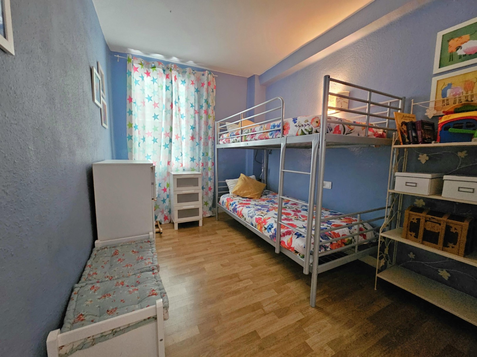 Slaapkamer met een metalen stapelbed, parketvloer en blauwe muren. De kamer is voorzien van witte opbergmeubels en een raam met gordijnen met sterrenprint.