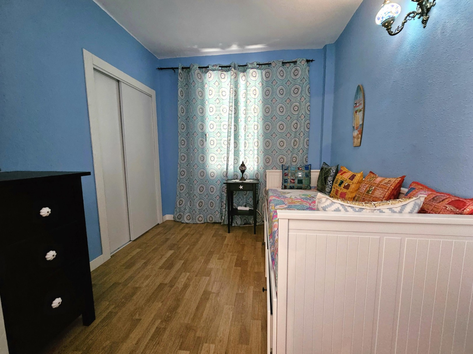 Slaapkamer met lichtblauwe muren, parketvloer en een witte inbouwkast. Voorzien van een wit uitschuifbed met kleurrijke kussens en een donkere ladekast.
