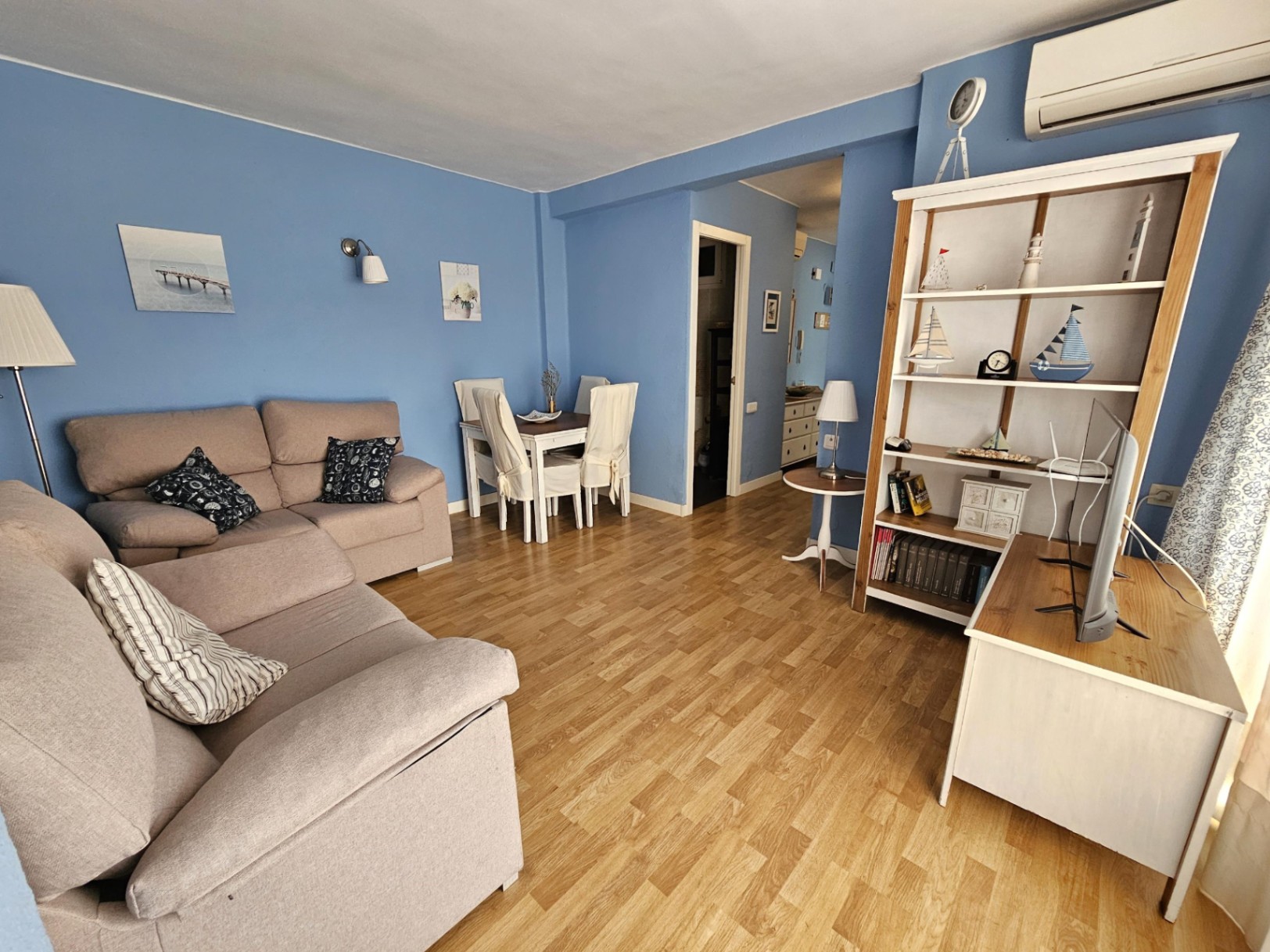 Woon- en eetkamer met blauwe muren, parketvloer en een beige bank. De ruimte is voorzien van een witte houten stellingkast en een eethoek voor vier personen.