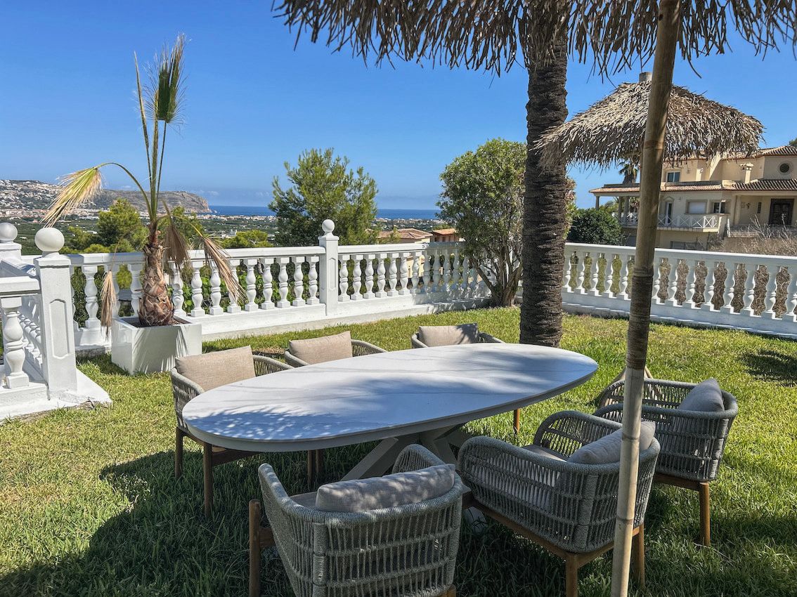 Buiten-eethoek op een gazon met een ovale tafel, gevlochten stoelen en een witte balustrade met panoramisch uitzicht op de vallei en de zee.