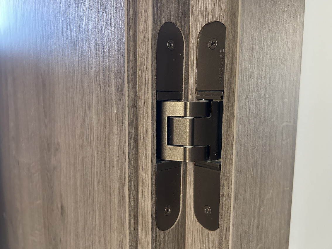 Detail van een hoogwaardig onzichtbaar deurscharnier in bronskleurige afwerking, gemonteerd in een houten kozijn als onderdeel van de kwalitatieve renovatie in 2025.