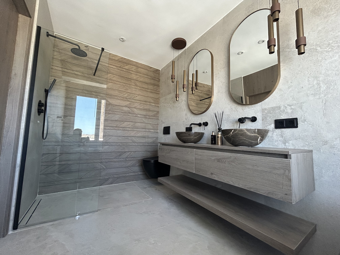 Moderne badkamer met dubbele stenen wastafel, zwarte kranen, ovale spiegels en een inloopdouche met houtlook tegelwand.