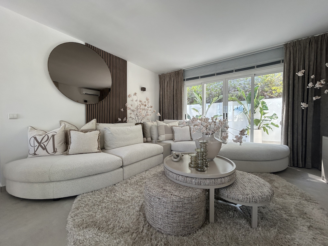 Moderne woonkamer met een gebogen neutrale bank, houten lattenwand met ronde spiegel en grote glazen schuifdeuren die toegang bieden tot het terras.