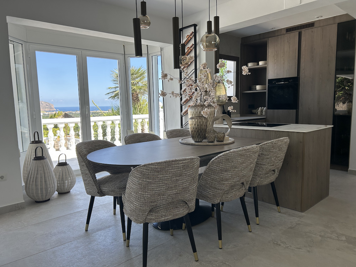 Open eetruimte met ovale tafel en moderne keuken. Grote raampartijen zorgen voor veel lichtinval en bieden een panoramisch uitzicht op de zee.