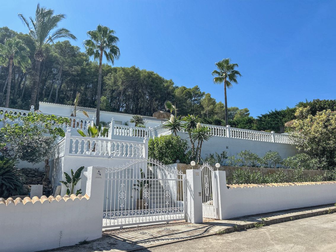 Witte gevel met meerdere niveaus, klassieke balustrades, een smeedijzeren toegangspoort en mediterrane beplanting met palmbomen.