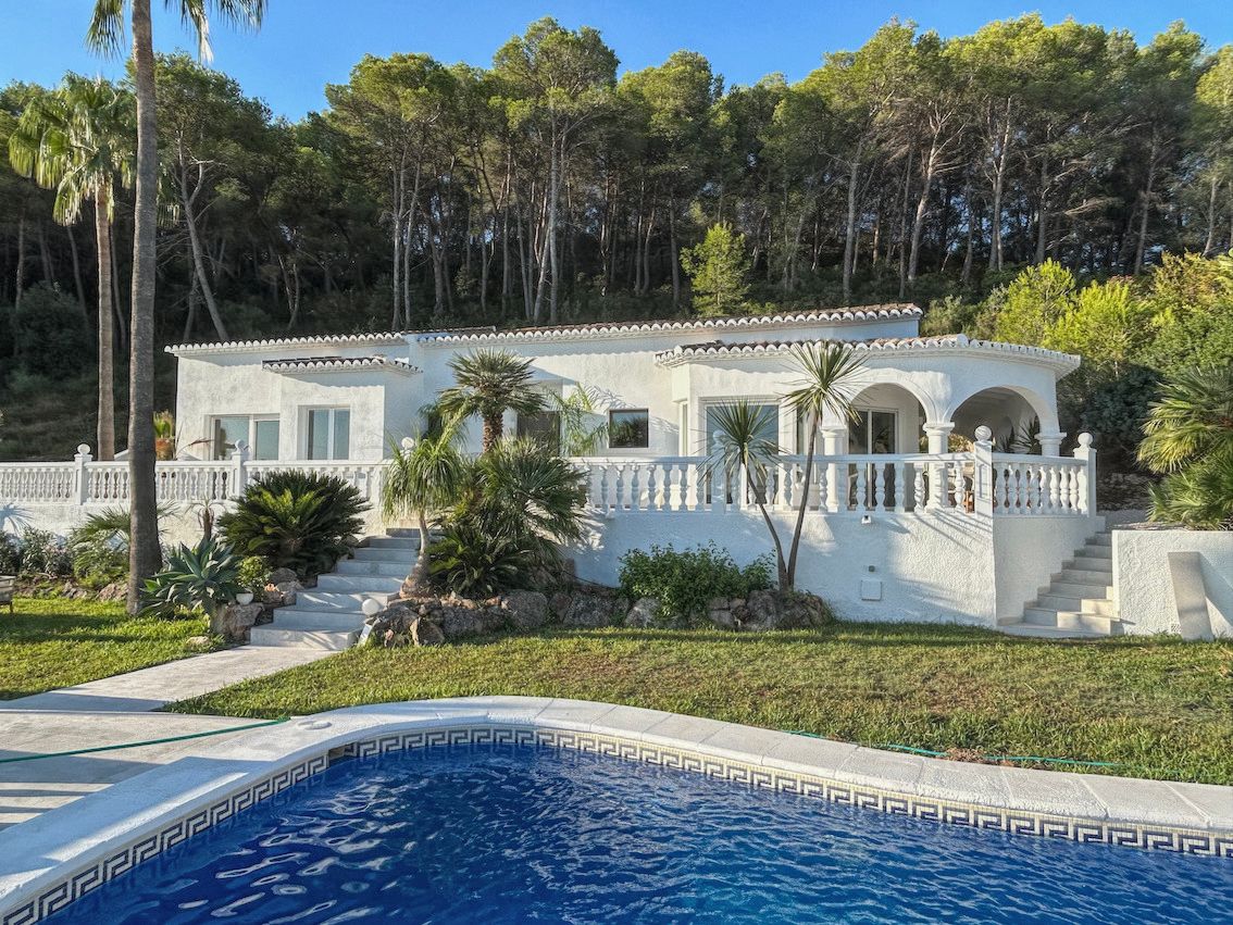 Witte villa met een terras met balustrades, mediterrane tuin en zwembad op de voorgrond, gelegen tegen een bosrijke achtergrond van pijnbomen.
