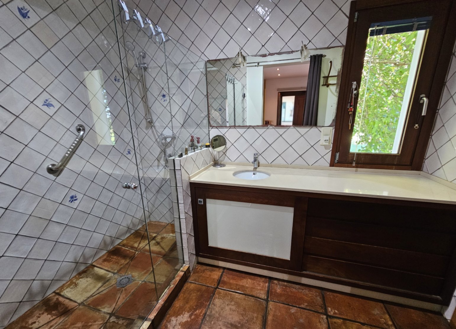 Badkamer met glazen douche, handbeschilderde wandtegels en rustieke kleivloeren. Voorzien van een donkerhouten wastafelmeubel en uitzicht op de tuin.