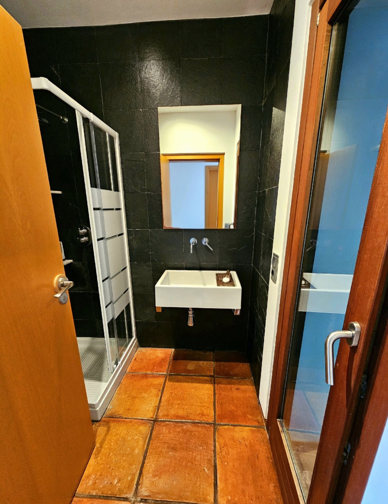 Badkamer met handgemaakte terracotta vloertegels, donkere natuurstenen wanden, een zwevende wastafel en een glazen douchecabine.