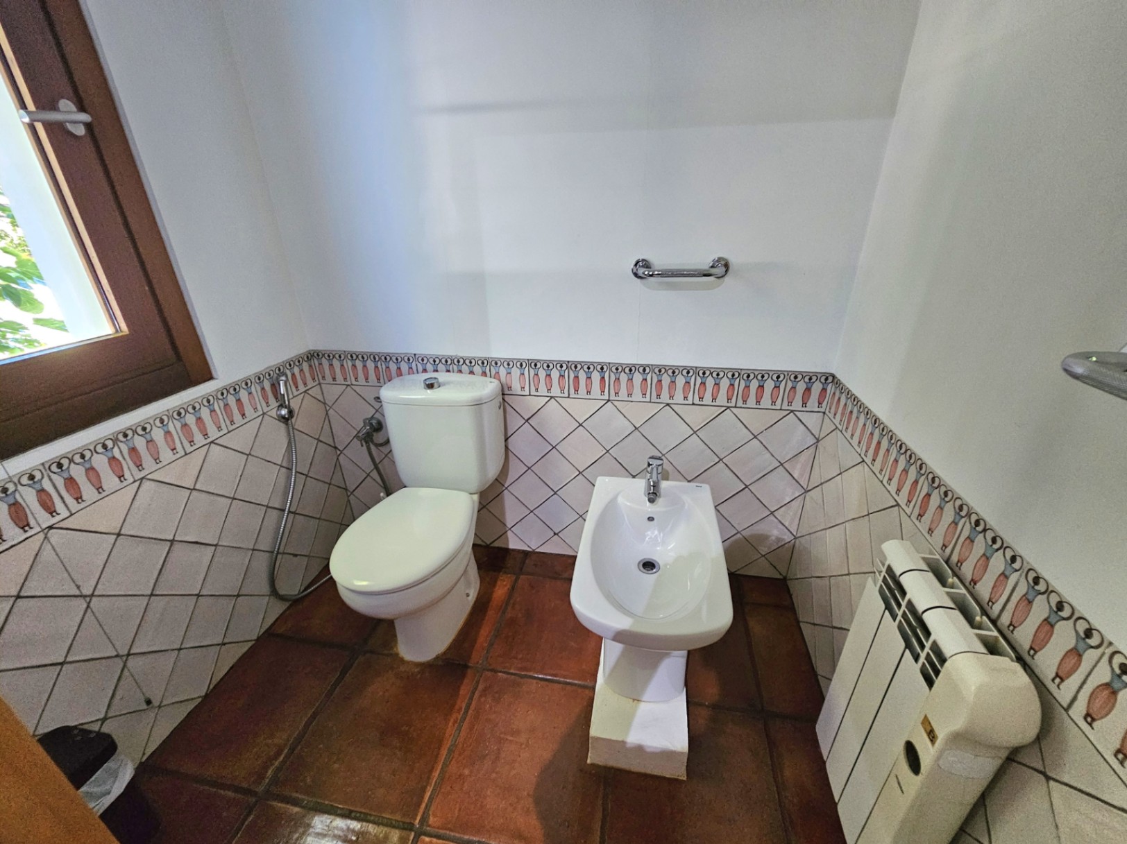 Badkamergedeelte met toilet en bidet, voorzien van handgemaakte terracotta vloeren en wandtegels met een handgeschilderde sierrand en houten kozijn.