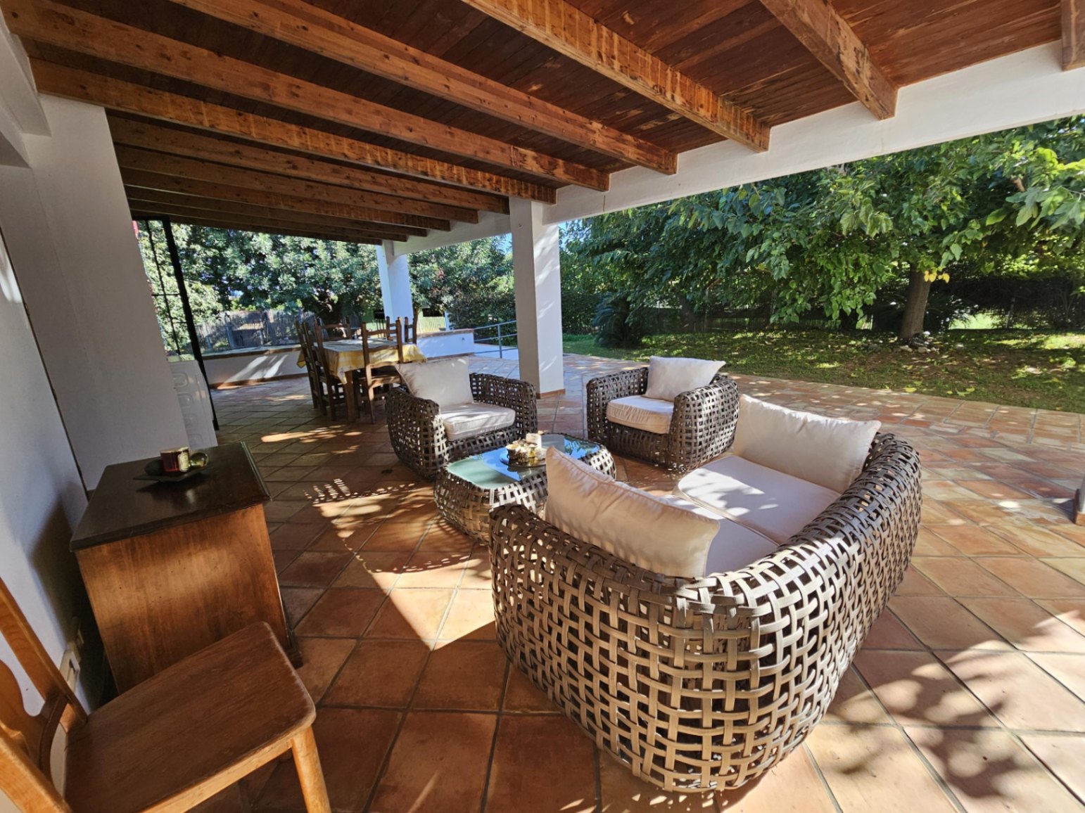Ruim overdekt terras met houten balkenplafond, handgemaakte terracotta tegels en gevlochten loungemeubels grenzend aan de volgroeide tuin.
